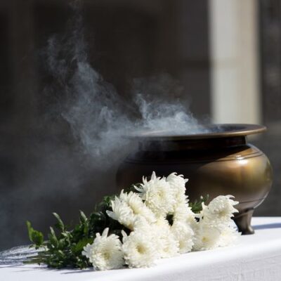 Une image à l'ambiance recueillie montrant un pot à encens en bronze tripode fumant, placé sur une table recouverte d'un drap blanc. Un bouquet de chrysanthèmes blancs est disposé devant le pot. De la fumée blanche s'échappe de l'encensoir sur un fond sombre.