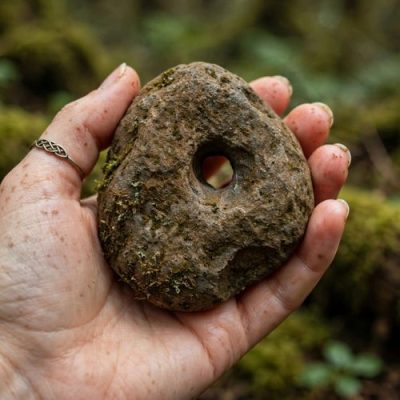 Gros plan sur une pierre de sorcière naturelle et percée en son centre, trouvée au bord d'une rivière, posée sur de la mousse verte comme talisman de protection ésotérique.