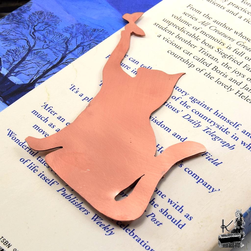 Marque-page chat en cuivre découpé à la main, posé sur un livre ancien près d'une bougie, accessoire de lecture féérique.