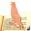 Marque-page corbeau en cuivre découpé à la main, posé sur un livre ancien près d'une bougie, accessoire de lecture féérique.