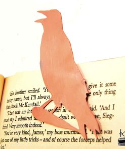 Marque-page corbeau en cuivre découpé à la main, posé sur un livre ancien près d'une bougie, accessoire de lecture féérique.